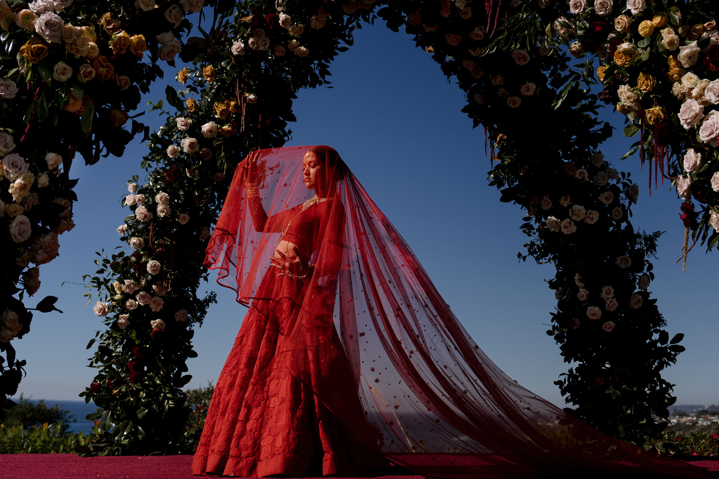Bride in red lehenga and matching red wedding veil standing under opulent floral arbor photographed by Matei Horvath Photography