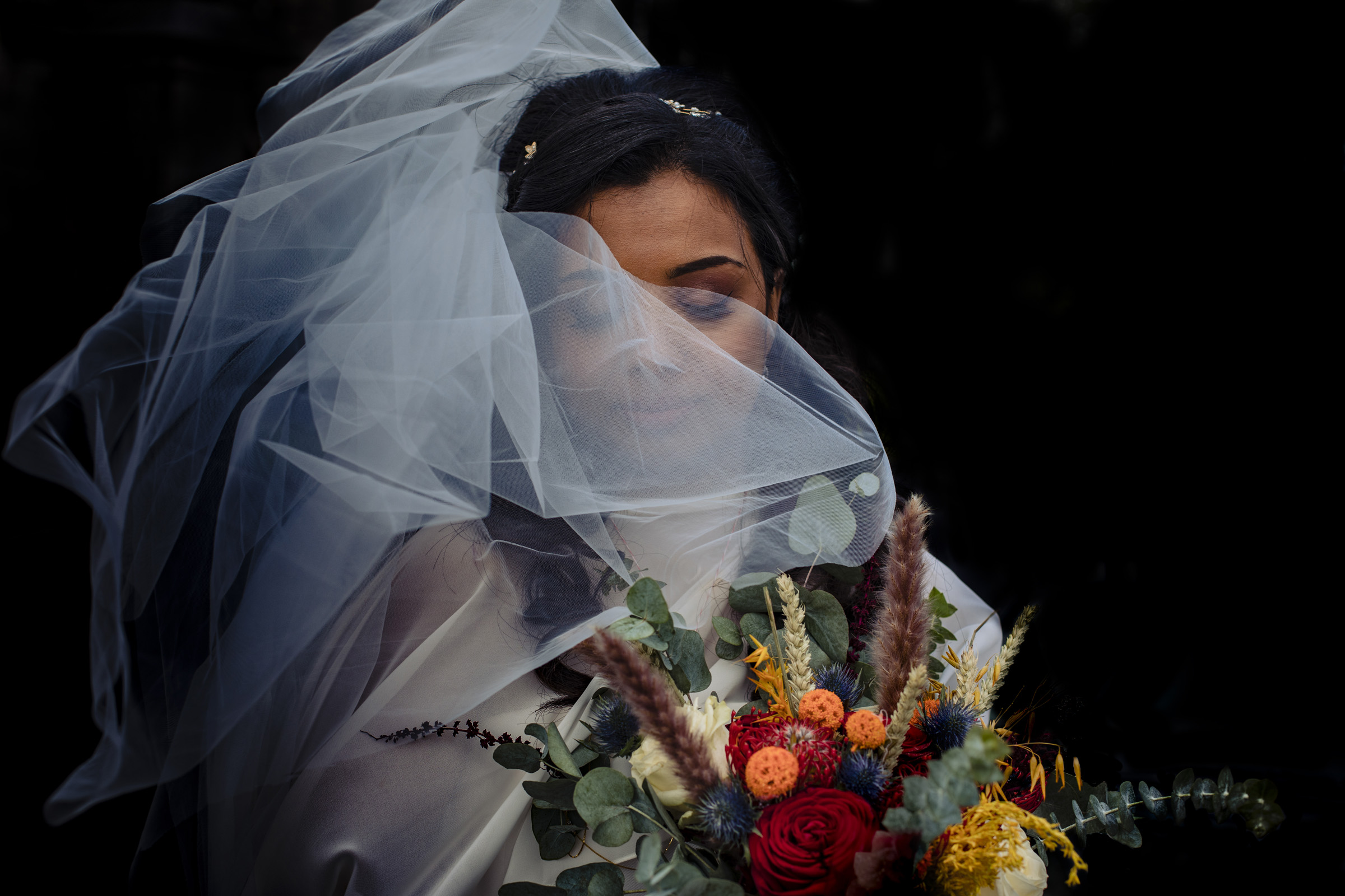 Bride holding colorful bouquet while wrapped in layers of tulle wedding veil for a romantic close-up by Stefano Ferrier