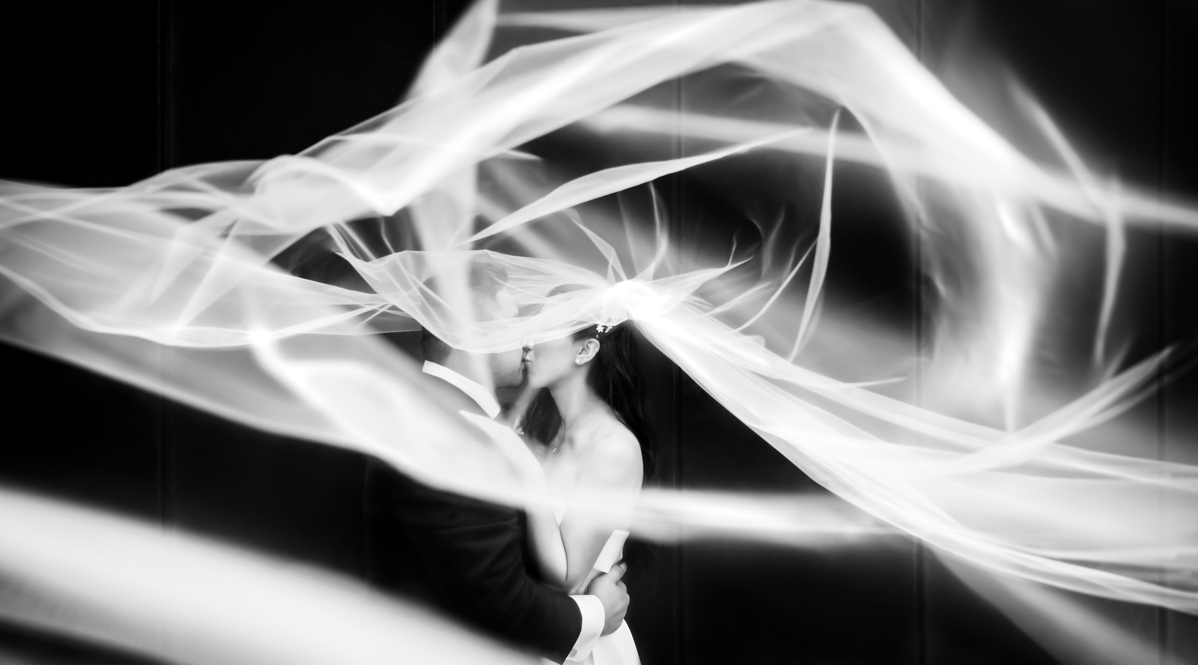 Couple kissing beneath a dramatically twirling wedding veil in black and white portrait by Dezine by Mauro