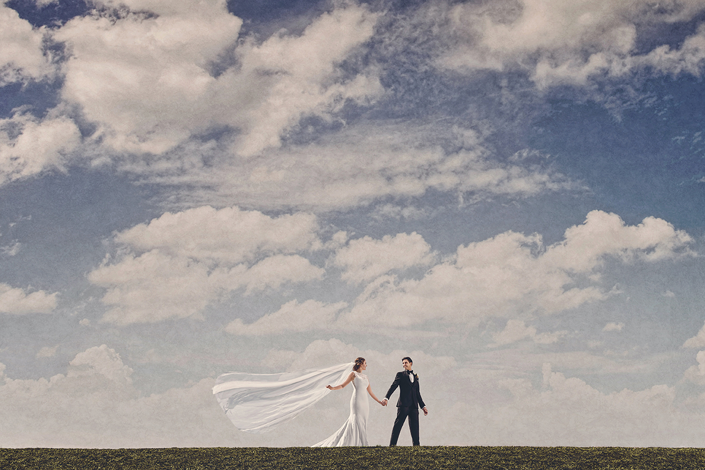 Bride and groom walking under vast blue sky with long flowing wedding veil trailing behind in landscape portrait by Gary Evans