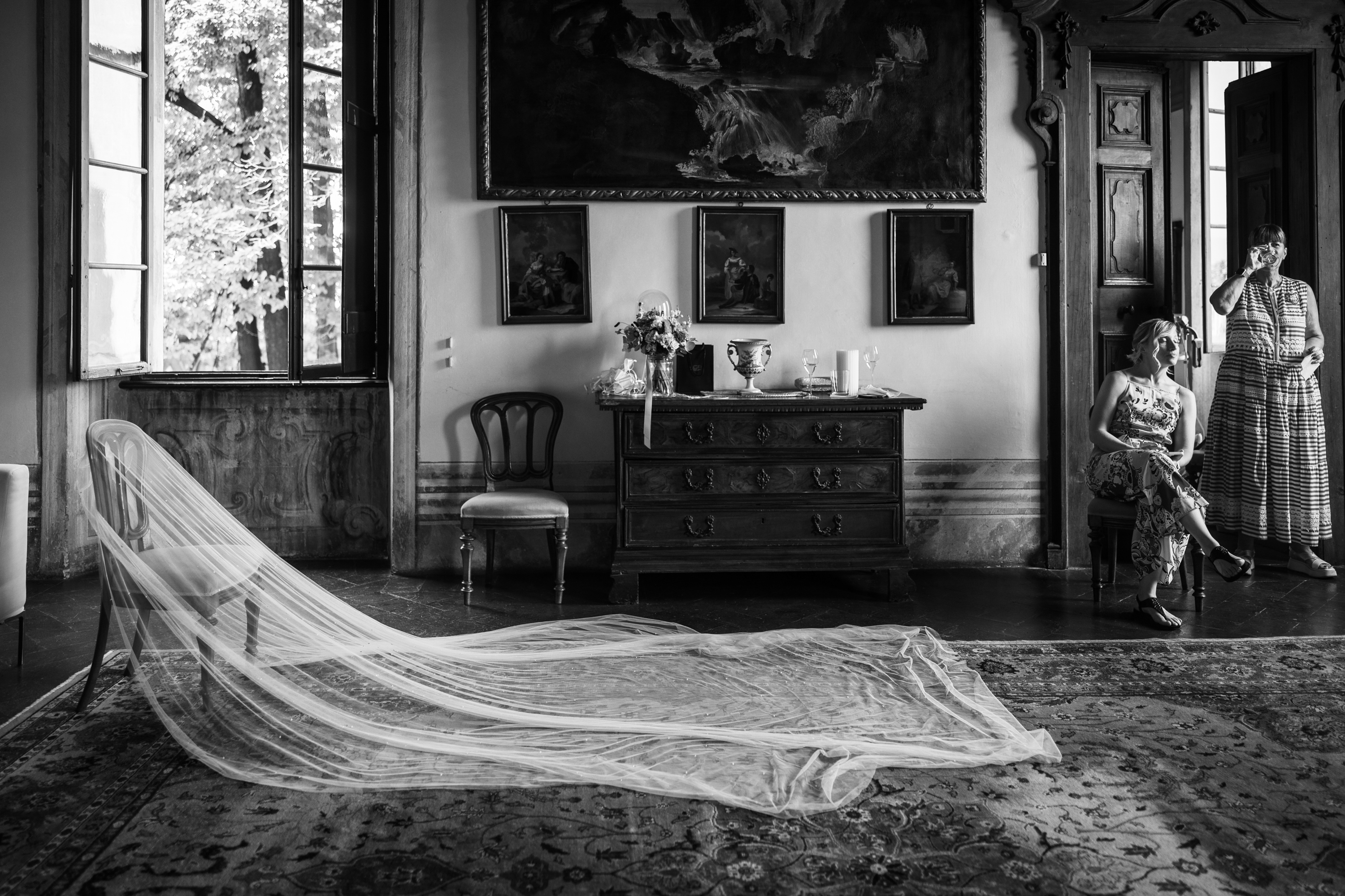 Long wedding veil draped over a chair in an elegant room with family members nearby photographed by Roberto Ricca Photo & Video