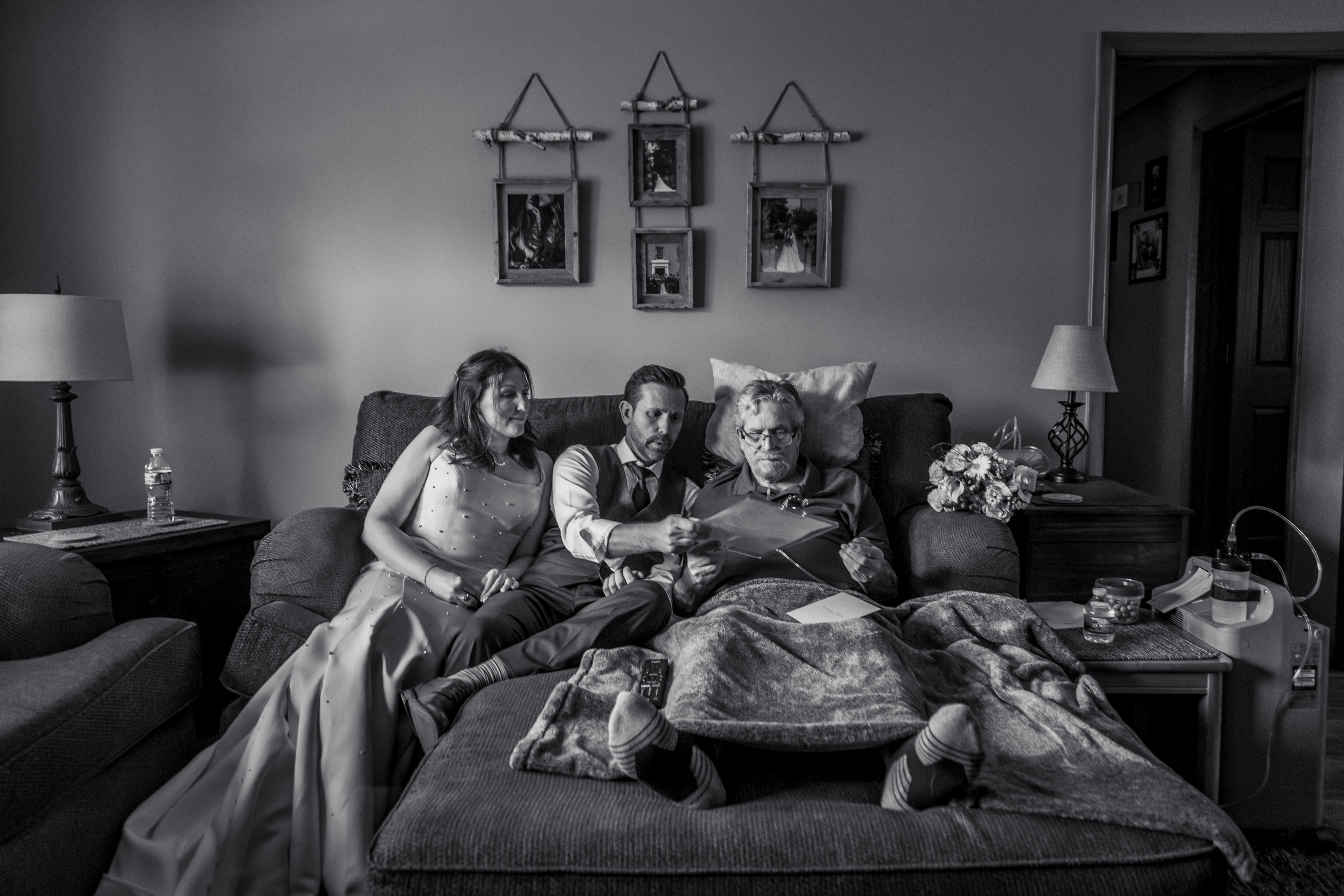 Black and white photo of couple showing wedding certificate to dad in bed by Amber Henry Photography