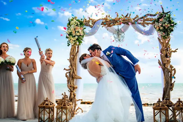 Bride and groom kiss at ceremony against blue sky and confetti by Michael Freas, North Carolina
