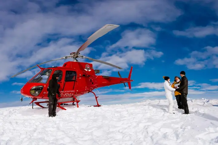 couple-on-snow-with-red-helicopter-under-blue-skies-pemberton-canada-photo-by-david-sherry-photography