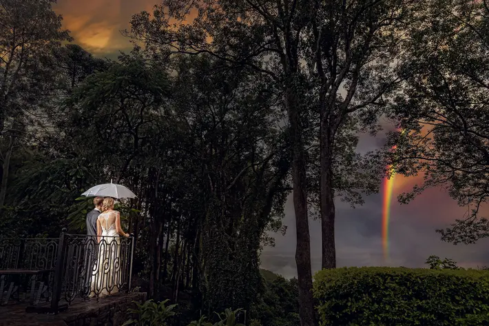 Couple under umbrella at rainy day wedding with rainbow, Ben Connelly, Australia