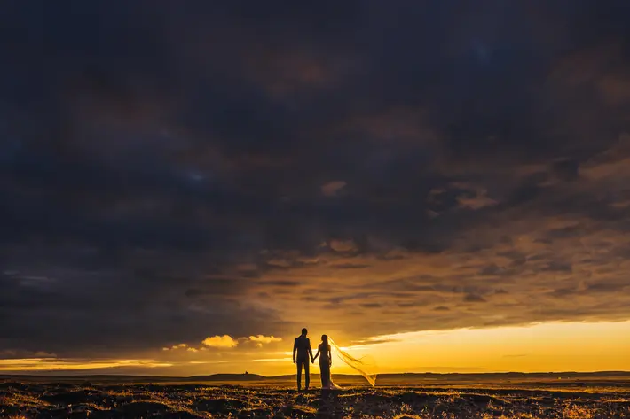 Bride and groom silhouetted under dark cloudy sunset photographed by Liam and Bee