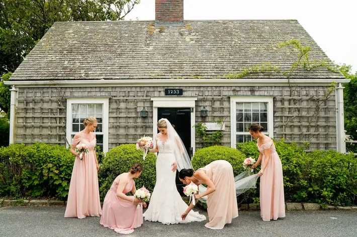Bridemaids in pink arrange bride's gown outside small cottage Kate McElwee Photography